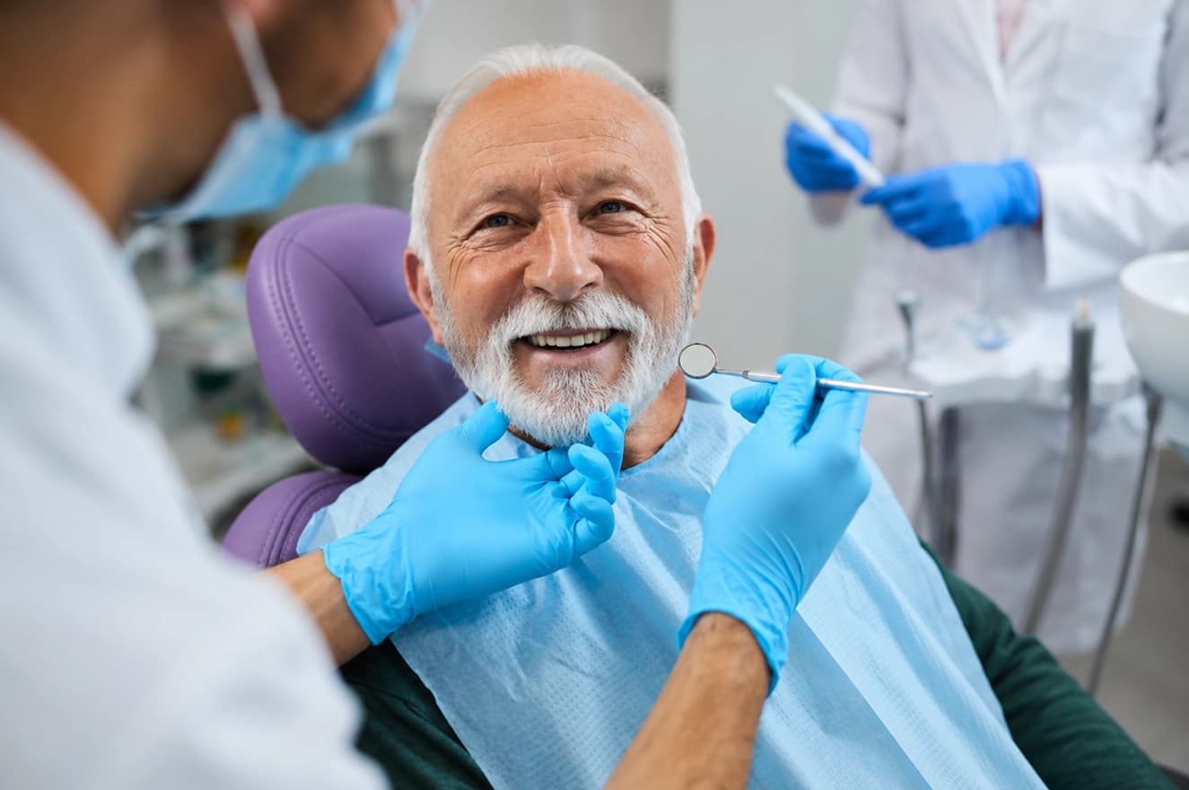 An older patient smiling in the chair while his dentist checks his teeth